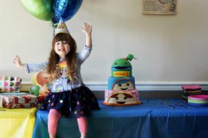 girl sitting on a table with a party hat next to a custom cake